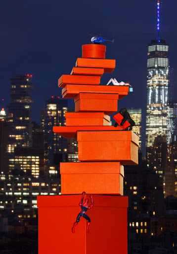 A whimsical nighttime scene features a tower of orange gift boxes against a city skyline, with miniature figurines and a toy helicopter.