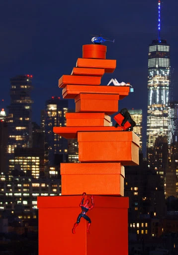 A whimsical nighttime scene features a tower of orange gift boxes against a city skyline, with miniature figurines and a toy helicopter.