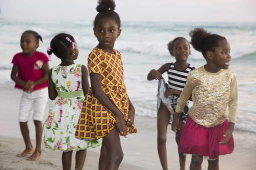 Five young black girls in colorful dresses playing on a sandy beach near ocean waves, captured in natural daylight.