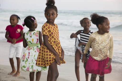 Five young black girls in colorful dresses playing on a sandy beach near ocean waves, captured in natural daylight.