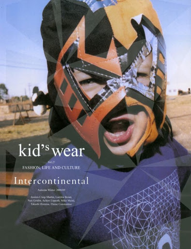 Child in lucha libre mask wearing a t-shirt posing in front of a magazine cover, outside on grass on a sunny day.
