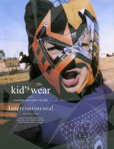 Child in lucha libre mask wearing a t-shirt posing in front of a magazine cover, outside on grass on a sunny day.