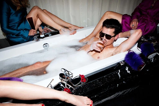 Glamorous scene features a man in a bubble bath with sunglasses, surrounded by three women's legs, shoes, and cocktail glasses.