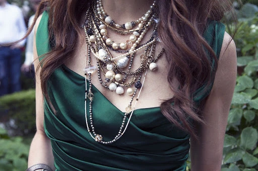 A woman in a green dress wears multiple strands of necklaces with pearls, beads, and crystals outdoors with green foliage visible.