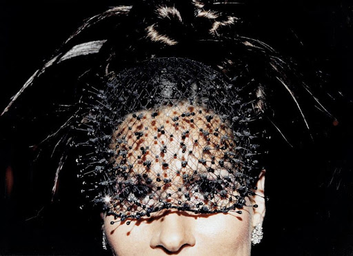 Close-up portrait of a female with beaded veil and feathered headpiece against a dark background, showcasing high-fashion and elegance.