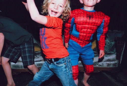 Three smiling children play indoors, one in a red Spider-Man costume, another in a red shirt and jeans, having fun with expressive body language.