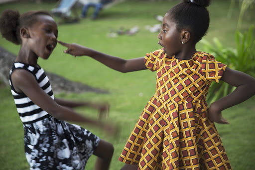 Two African girls playfully interacting outdoors, one pointing a finger at the other's face in a grassy, natural environment.
