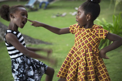 Two African girls playfully interacting outdoors, one pointing a finger at the other's face in a grassy, natural environment.