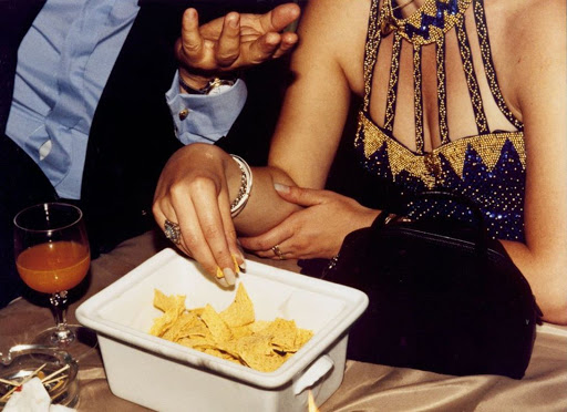Indoor social event at night: woman in beaded dress takes a chip. Nearby man gestures, orange drink, purse. Low-light indoor photography.