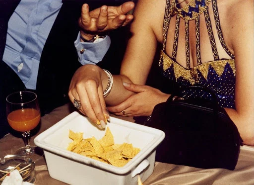 Indoor social event at night: woman in beaded dress takes a chip. Nearby man gestures, orange drink, purse. Low-light indoor photography.