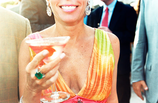 Close-up of a woman holding a pink drink in a stemmed glass, wearing a colorful dress and emerald ring, with people in the background.