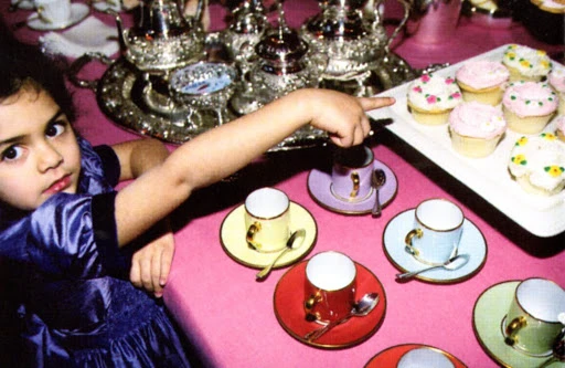 A young child in a blue dress points at cupcakes during an indoor tea party, surrounded by tea sets and a pink tablecloth.