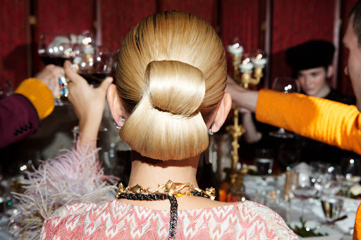Blonde updo hairstyle, back view of an adult woman wearing a pink and white dress, with people at a dinner party in the background.