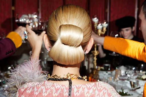Blonde updo hairstyle, back view of an adult woman wearing a pink and white dress, with people at a dinner party in the background.