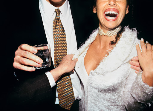 A woman in a white jacket and pearl choker smiles with a man in a suit holding a drink, standing against a dark background.