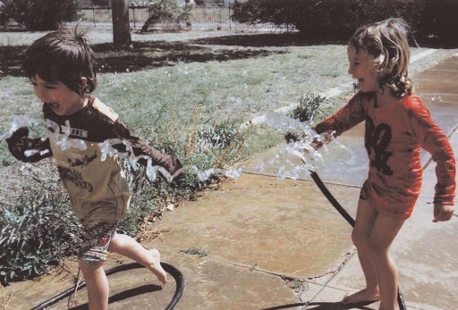Two young children gleefully play with water from a garden hose on a sunny day in a yard, expressing joy and laughter.