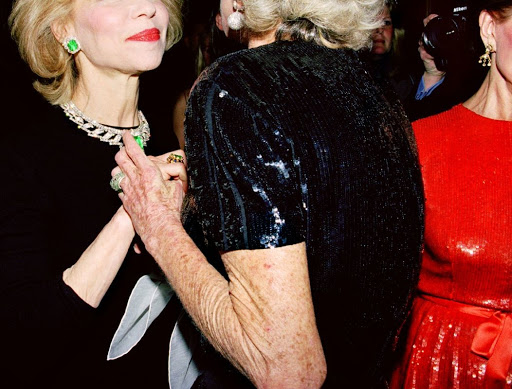 Three women dressed in formal attire attend an indoor social event. Emerald jewelry, shiny fabrics, and camera flash are visible.
