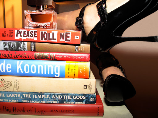 Close-up of black high heels resting on a stacked pile of books next to a perfume bottle by photographer Jessica Craig-Martin.