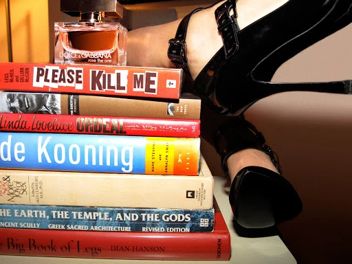 Close-up of black high heels resting on a stacked pile of books next to a perfume bottle by photographer Jessica Craig-Martin.