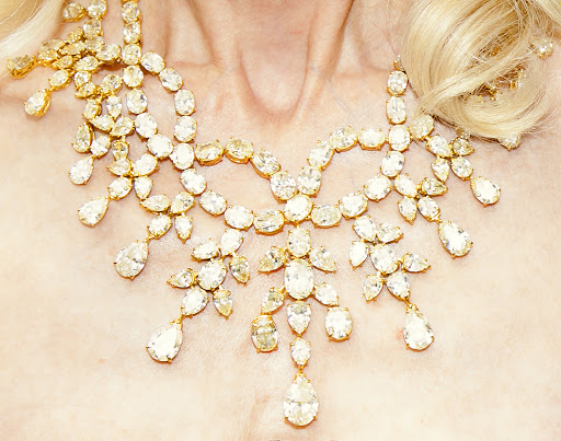 Close-up of a woman's neck and upper chest adorned with a large, ornate gold necklace with clear gemstone pendants and teardrop stones.