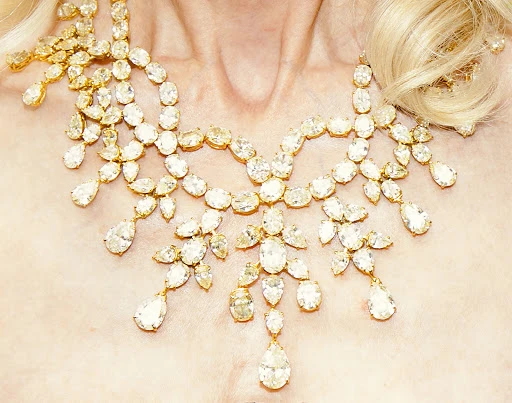 Close-up of a woman's neck and upper chest adorned with a large, ornate gold necklace with clear gemstone pendants and teardrop stones.