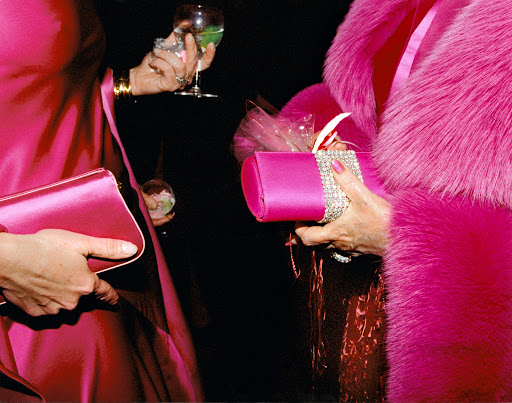 Two women in bright pink dresses holding a cocktail and a wrapped gift with shiny accents at an event with a dark background.