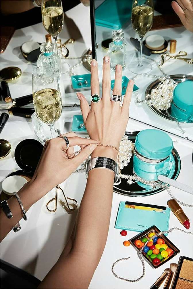 Overhead shot showcasing a woman's hands adorned with jewelry from Tiffany & Co., surrounded by champagne, candy, and various beauty products.