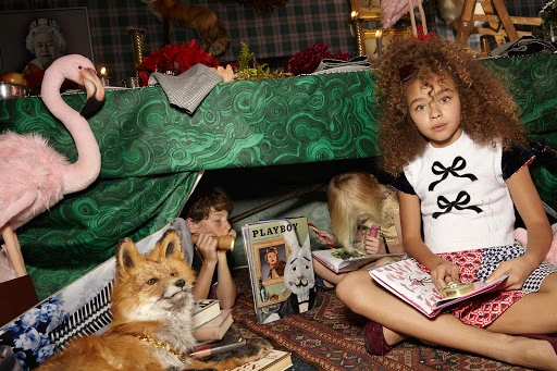 Young girl with curly hair sits cross-legged reading a book inside a tent-like structure with other kids, taxidermy fox, flamingo.