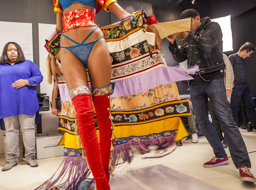 Fashion shoot backstage with model in lingerie, floral skirt, red thigh-high boots, and photographer using a makeshift cardboard lens.