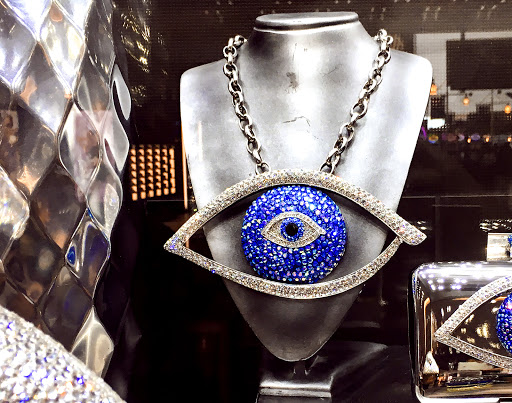 Necklace and a handbag with blue jeweled eye design on a mannequin against a black background with textured metallic accents.