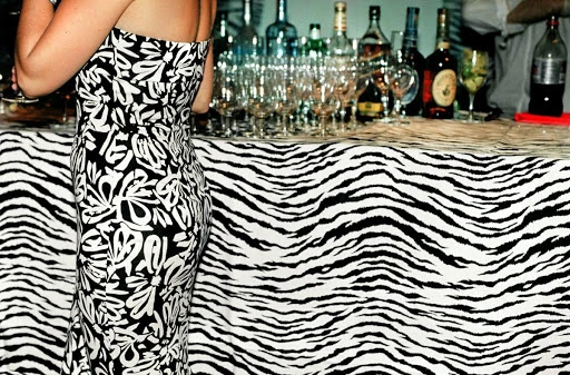A woman in a black-and-white dress stands near a table covered with a zebra-print cloth, bottles, and glassware.