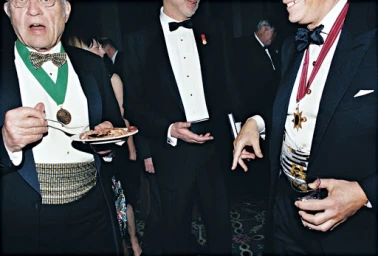 Group of men in black tie tuxedos at a formal event wearing medals, sashes, and cummerbunds while eating and holding drinks