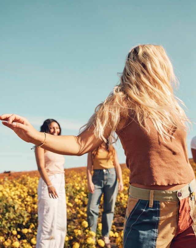 Several women in field of yellow flowers under a blue sky, with one woman in brown crop top and colorful pants prominent in the foreground.