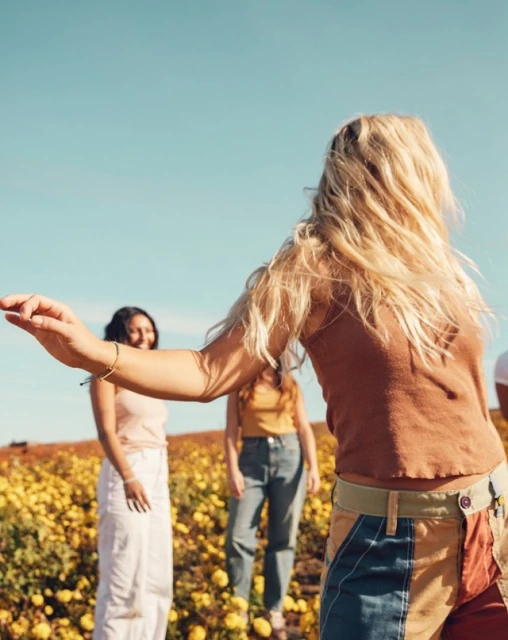 Several women in field of yellow flowers under a blue sky, with one woman in brown crop top and colorful pants prominent in the foreground.