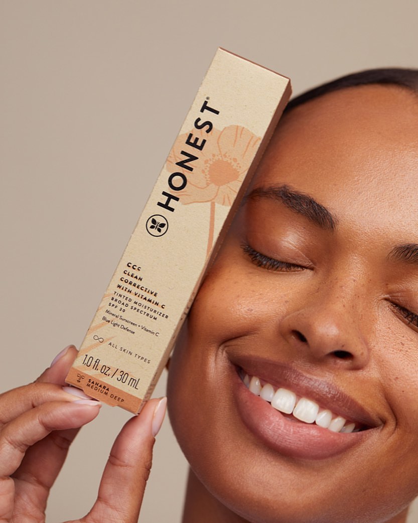 Tan-skinned woman smiles, holding a Honest Company CCC tinted moisturizer tube in studio light against a beige backdrop. Close-up.