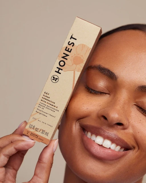 Tan-skinned woman smiles, holding a Honest Company CCC tinted moisturizer tube in studio light against a beige backdrop. Close-up.