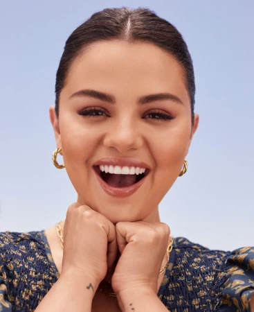 Close-up portrait of Selena Gomez smiling broadly with hands under chin against a pale blue sky background