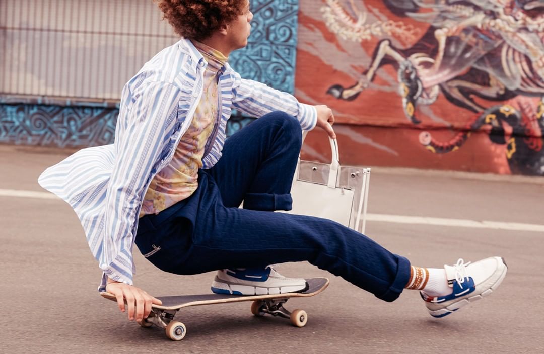 A person rides a skateboard down an asphalt street with a clear tote bag, wearing jeans and a striped shirt, with graffiti wall background.