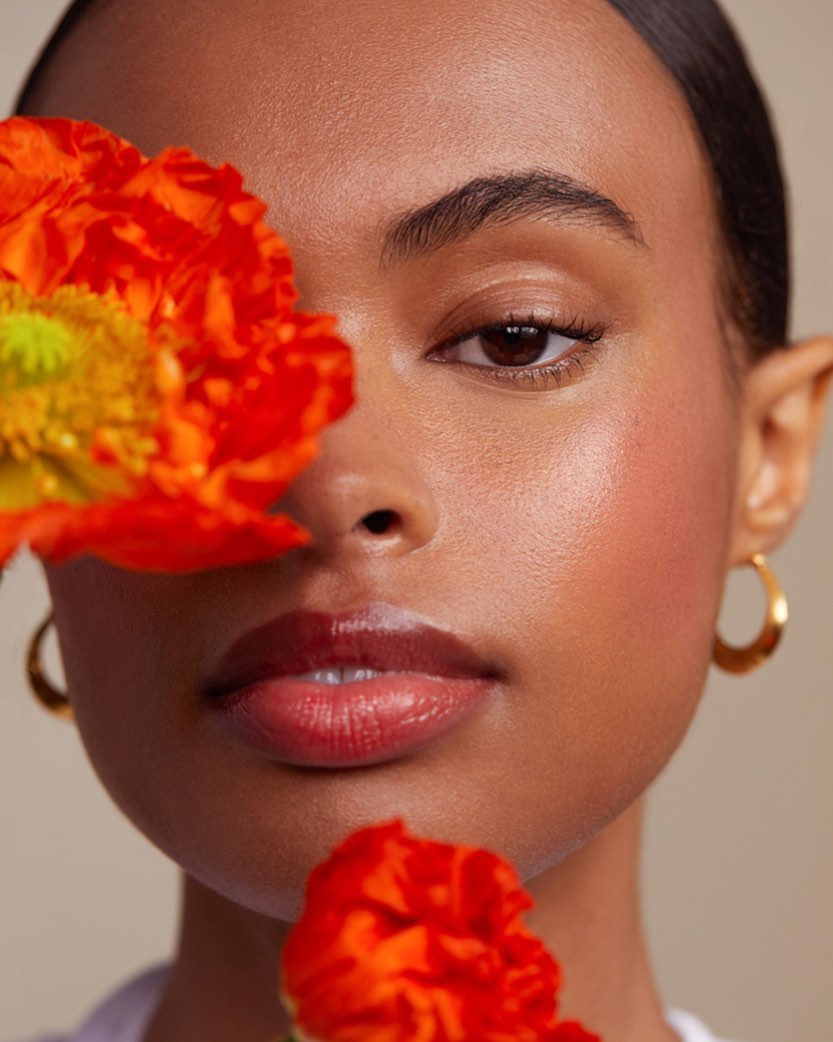 Close-up portrait of a woman with dark hair and hoop earrings, posing with orange flowers near her face in a beauty setting.