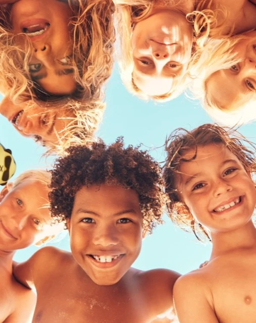 Group of smiling children looking down into the camera against a blue sky backdrop, showcasing friendship, togetherness, and summer fun.