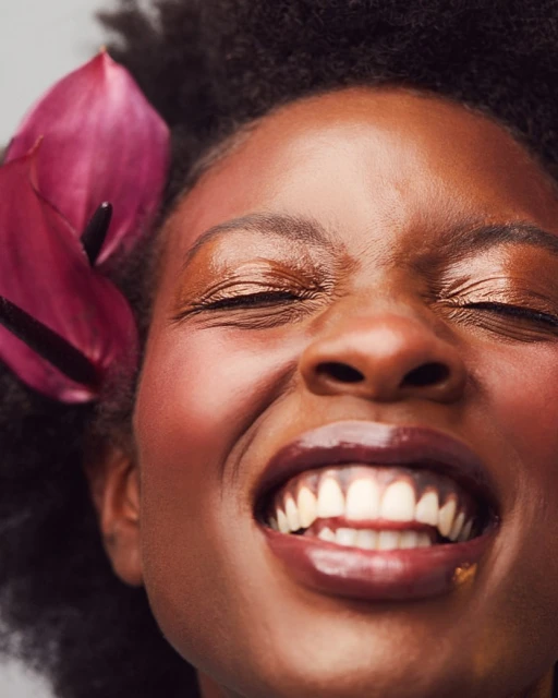 Close-up portrait of a smiling African American woman with closed eyes and dark curly hair, accentuated by a vibrant pink calla lily.