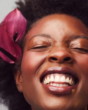 Close-up portrait of a smiling African American woman with closed eyes and dark curly hair, accentuated by a vibrant pink calla lily.