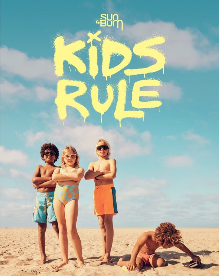 Group of four kids on beach wearing swimwear under bright blue sky with clouds and a yellow brand logo and text design above.