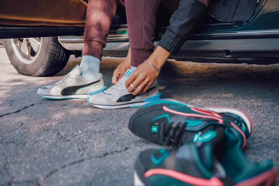 Person sitting in car doorway ties gray and black Puma sneaker; a pair of sneakers are on the cracked asphalt ground.