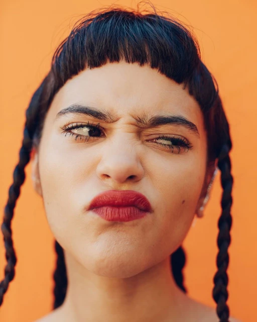 Close-up portrait of a woman with braided hair, red lipstick, and a slightly cross-eyed expression against an orange backdrop.