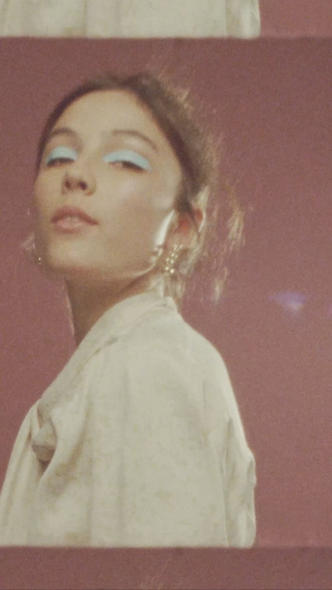 Woman with light skin wearing light blue eyeshadow and a beige jacket posing indoors against a pink wall with natural lighting.