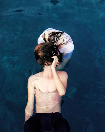 High-angle view of two people in the blue water of a pool, one fair-skinned male torso, and a female in a white top with wet dark hair.