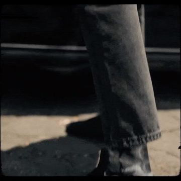 A person walks on a sidewalk wearing denim jeans and dark shoes with a dark vehicle parked in the background on a city street.