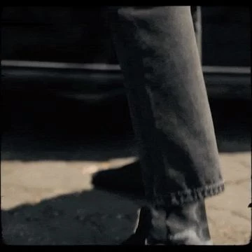 A person walks on a sidewalk wearing denim jeans and dark shoes with a dark vehicle parked in the background on a city street.