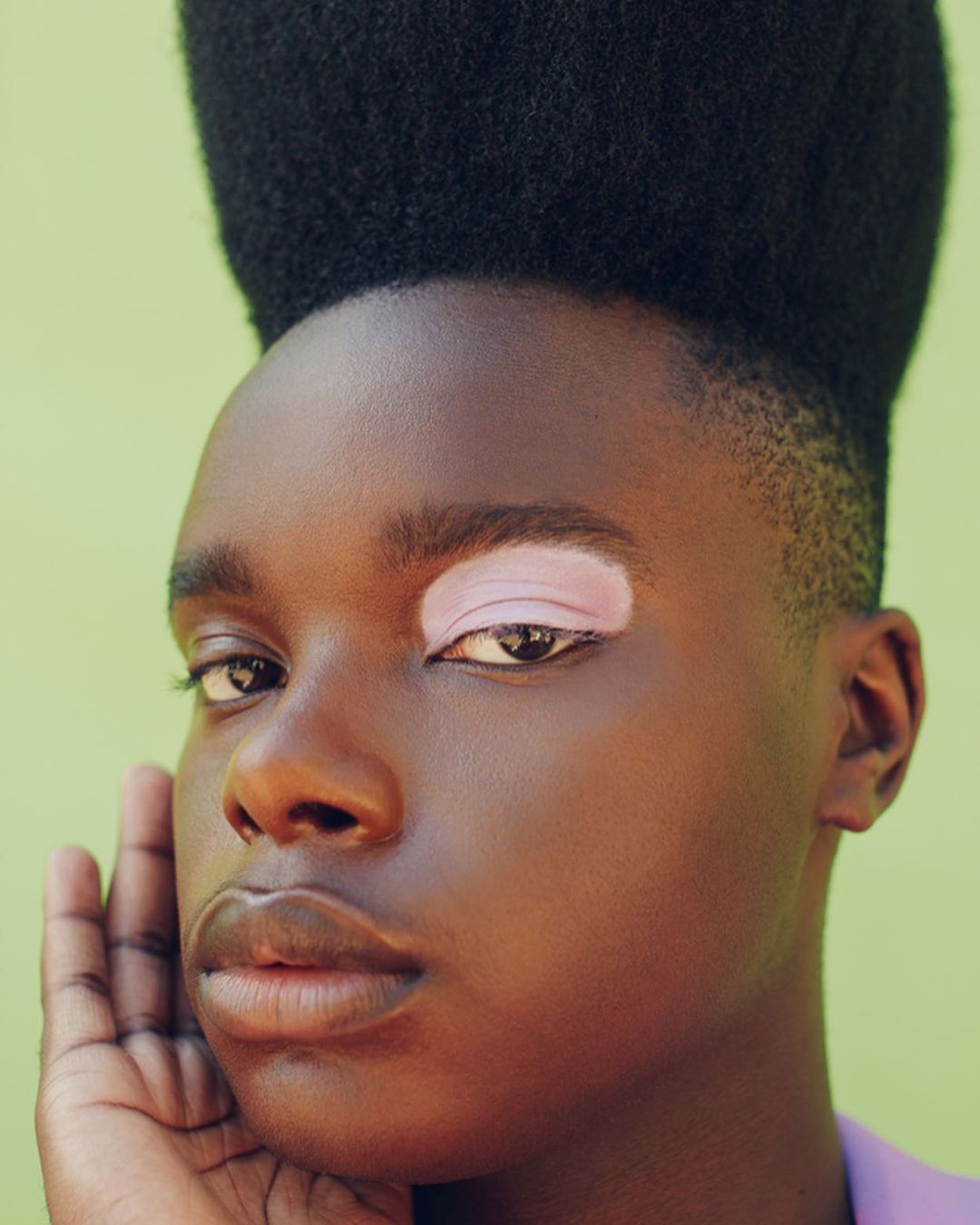 Close-up portrait of a young, dark-skinned adult with a high-top fade hairstyle against a light green background in a photo studio setting.